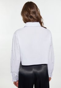 Blusa bianca accorciata con colletto a punta e maniche lunghe, caratterizzata da decorazioni metalliche sulle spalle. Tessuto liscio e leggero.