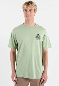 T-shirt en coton vert clair à manches courtes, col rond, avec un logo graphique noir circulaire sur le côté gauche de la poitrine. Coupe lisse et décontractée.
