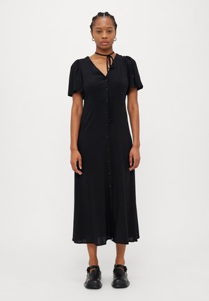 Black mid-length dress with puff sleeves, V-neck, button front, and a tie detail. Worn with black slip-on shoes, smooth fabric texture.