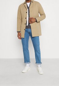 Beige bomullskappa med knappstängning, tillsammans med en grå kofta och blå jeans med uppvikta ärmar. Vita sneakers fulländar looken.