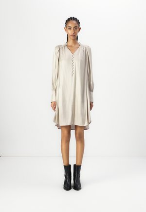 Beige, pitkähihaine mekko, jossa on V-aukko ja etunappiyksityiskohtia. Pehmeä kangas, rento istuvuus, päättyy polvien ylle, yhdistetty mustiin saappaisiin.