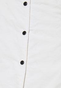 Camisa blanca con una textura suave, que presenta botones negros a lo largo del centro y costuras visibles. Diseño simple, sin patrones ni acentos.