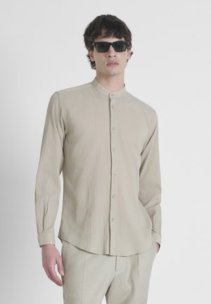 Camicia beige a maniche lunghe con colletto alla cinese, chiusura a bottoni, tessuto strutturato e orlo curvo, abbinata a occhiali da sole scuri.