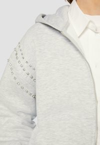 Grauer Reißverschluss-Hoodie aus strukturiertem Stoff mit dekorativen Strasssteinen entlang der Schulternaht. Kapuze und Reißverschlussdetails sichtbar.