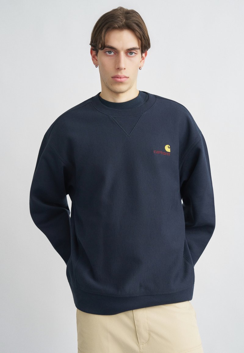 Πανωφόρι φούτερ σε ναυτικό μπλε χρώμα, κατασκευασμένο από βαμβακερό μείγμα, με ραγκλάν μανίκια και μικρό κίτρινο λογότυπο με το "carhartt" κεντημένο με κόκκινο στο στήθος.
