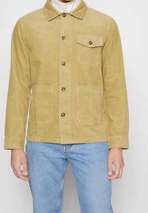 Mężczyzna w musztardowo-żółtej kurtce sztruksowej z przodem na guziki, trzema kieszeniami, w połączeniu z jasnoniebieskimi dżinsami i białym golfem.