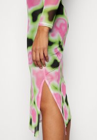Robe à manches longues avec un motif tie-dye en rose vif, vert et noir. Présente une fente sur le côté et un tissu doux et extensible pour le confort.