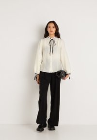 Λευκή μπλούζα με ψηλό γιακά και μαύρη κορδέλα, συνδυασμένη με μαύρα παντελόνια. Η μπλούζα διαθέτει μακριά, φουσκωτά μανίκια και κουμπιά.