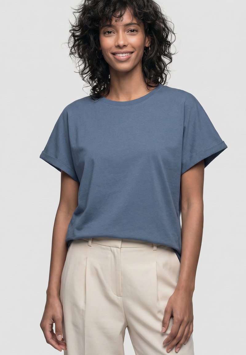 Femme souriante aux cheveux bouclés portant un t-shirt bleu aux manches retroussées et un pantalon beige taille haute devant un fond uni.
