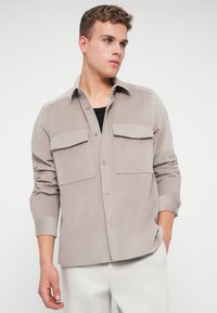 Camisa de corduroy color beige claro con dos bolsillos frontales, diseño de botones y mangas enrolladas. Textura suave y ajuste casual.