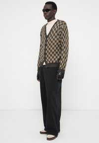 Cardigã xadrez em lã castanha e bege, com design em v, frente com botões, usado sobre uma blusa clara. Combinado com calças pretas riscas e luvas.