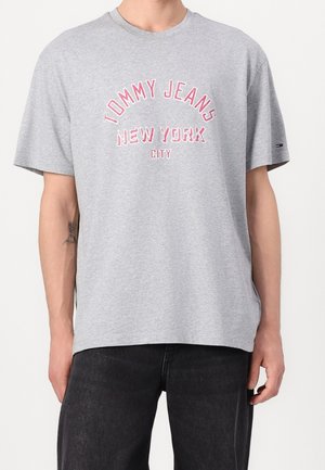 Jasnoszara koszulka z krótkim rękawem z różowym i białym tekstem "Tommy Jeans New York City", noszona przez męskiego modela w czarnych spodniach.