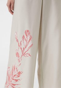Lichtbeige stoffen broek met een groot, textuurachtig bladmotief in koraal aan de zijkant, die een moderne en minimalistische stijl uitstraalt.