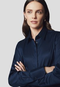 Granatowa koszula zapinana na guziki z gładkiego materiału, z klasycznym kołnierzykiem, długimi rękawami i mankietami zapinanymi na guziki. Dodatki obejmują kolczyki.