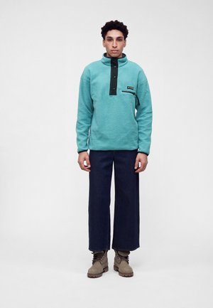 Jeune homme portant un pull polaire bleu sarcelle, un pantalon large foncé et des bottines beiges lacées, debout devant un fond uni.