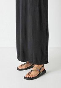 Schwarzes Maxikleid, das bis zur Mitte der Wade reicht, kombiniert mit flachen Sandalen mit lebhaften, mehrfarbigen Riemen und einer robusten schwarzen Sohle.