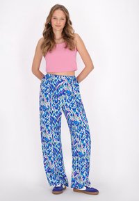 Rosa, kurz geschnittenes Tanktop und locker sitzende Hose mit einem abstrakten Muster in Blau, Grün und Pink. Das Model trägt blaue Turnschuhe.