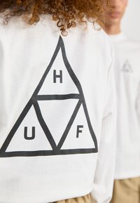 Hvit langermet skjorte med en stor svart trekantet grafikk med bokstavene "U", "H" og "F". Myk stoff og avslappet passform.