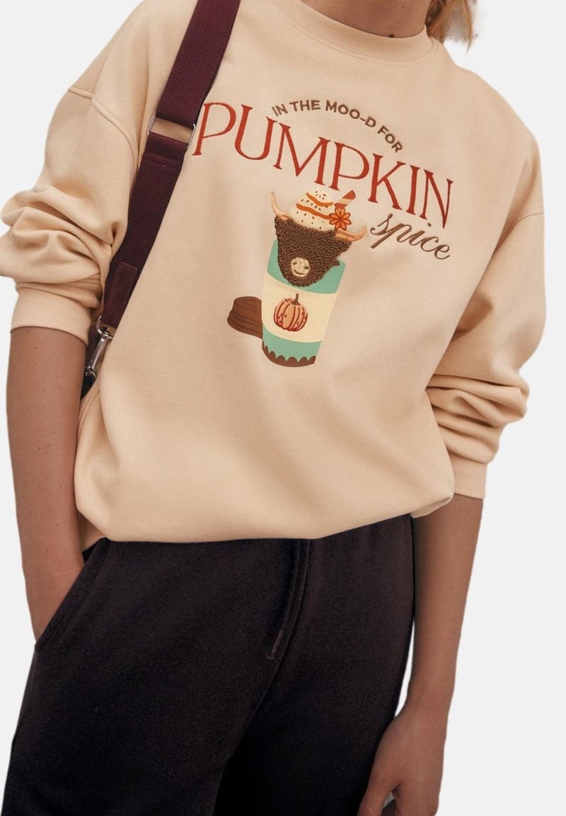Beige trui met grafisch ontwerp met een pumpkin spice drankje en dierenmotief. Tekst luidt "IN THE MOOD FOR PUMPKIN spice."