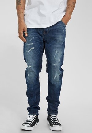 Uomo che indossa jeans blu consumati, t-shirt bianca, sneakers alte nere, con avambracci tatuati e anelli sulla mano sinistra.