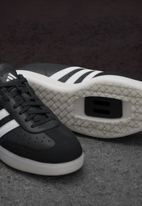 Czarne buty sportowe wykonane ze skóry, z perforowanymi detalami, z trzema białymi paskami, teksturowaną białą podeszwą i widocznym logo.