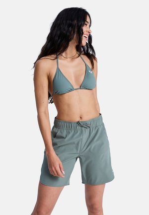 Kobieta z długimi ciemnymi włosami, ubrana w zielony top bikini i pasujące zielone szorty z troczkiem, uśmiechająca się i spoglądająca w bok.