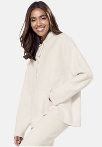 Bež velika kapuca s vezicami, sprednjim žepom, rebrastimi manšetami in robom. Mehka tkanina in sproščen kroj za udobje.