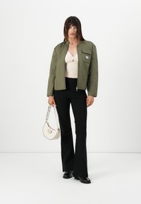 Giacca imbottita verde oliva con zip frontale e taschino sul petto, abbinata a un top chiaro e pantaloni neri svasati. Include una piccola borsa bianca.