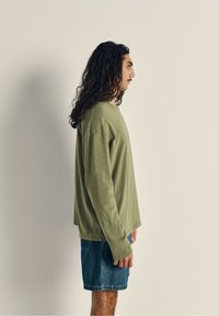 Oliven grøn langærmet skjorte med afslappet pasform og rund halsudskæring, parret med denimshorts. Teksturen ser blød og afslappet ud.