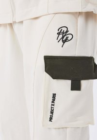 Pantalon cargo couleur crème avec une grande poche verte olive, logo brodé et tissu texturé. Présente des coutures doubles et un design minimaliste.