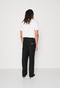 Zwarte cargobroek met meerdere zakken, recht model en een klein logopatch op de achterkant. Draagt met een witte t-shirt met korte mouwen.
