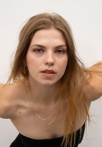 Goldene Ketten mit einer strukturierten Oberfläche, getragen über einem schwarzen trägerlosen Oberteil. Die Ohrringe sind einfache goldene Creolen, die den Look ergänzen.