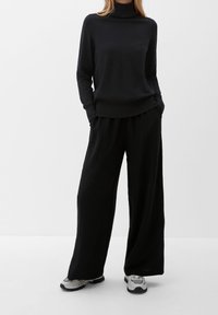 Pull noir à col montant avec manches longues, associé à un pantalon noir à jambes larges texturé et des baskets grises avec des accents noirs.