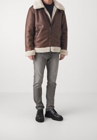 Giacca bomber in pelle marrone con colletto e polsini in shearling sintetico, abbinata a jeans grigi e scarpe nere stringate.