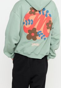 Mintgroene hoodie met een groot meerkleurig bloemmotief als patch. Voorzien van een zachte textuur en een capuchon met trekkoord, gecombineerd met zwarte broek.