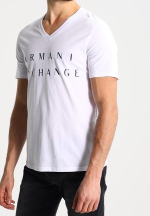 Homme portant un t-shirt blanc col en V Armani Exchange et un pantalon noir, debout avec les bras détendus devant un fond uni.