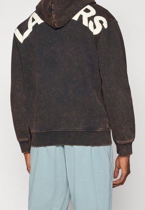 Person trägt einen ausgewaschenen schwarzen Hoodie mit großen weißen Buchstaben auf den Schultern und hellblauen Hosen, die von der Kamera weg zeigt.