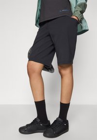 Shorts noirs en tissu extensible, dotés de poches plates, d'une coupe ajustée, portés avec des baskets noires et des chaussettes.