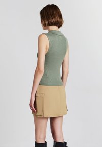 Donna di spalle con indosso un top verde a coste senza maniche, una mini gonna beige con tasche e stivali neri fino al ginocchio su uno sfondo neutro.
