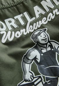 Grüner Stoff mit weißem Text, der "PORTLAND Workwear" über einer schwarz-weißen Illustration eines Arbeiters, der eine Farbkanne hält, spell.