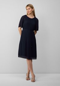 Robe plissée marine à manches courtes, décolleté rond, taille cintrée, jupe fluide et détail noué, associée à des sandales à talons nudes.