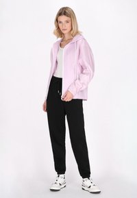 Giacca zip-up rosa chiaro con cappuccio regolabile, abbinata a pantaloni della tuta neri e sneakers bianche. Presenta lacci e una vestibilità strutturata.