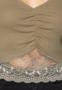 Brun ribbad tygblus med en rynkad mittsöm och blommig spetskant över svarta byxor.