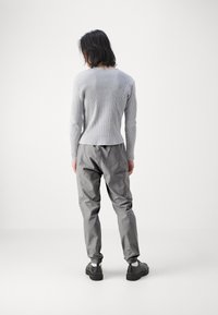 Grå, ribbad långärmad skjorta i kombination med lösa gråbyxor; vita manschetter synliga och svarta skor med tjock sula. Minimalistisk bakgrund.