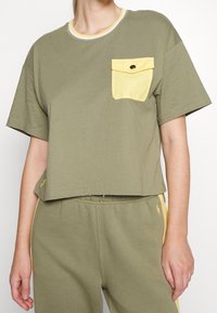 Olivově zelené zkrácené tričko vyrobené z měkké látky, s žlutou kapsou s detailem knoflíku a kontrastním obšitím výstřihu.