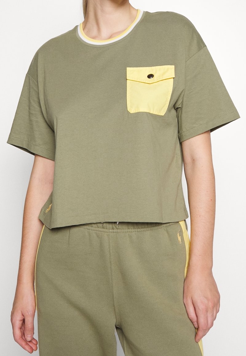 Olivově zelené zkrácené tričko vyrobené z měkké látky, s žlutou kapsou s detailem knoflíku a kontrastním obšitím výstřihu.