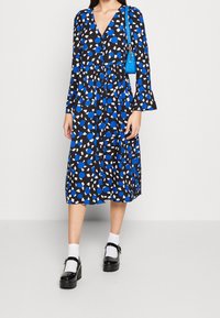 Abito nero con motivo astratto blu e bianco, scollo a V, maniche lunghe, polsini svasati, vita abbinata, accompagnato da una piccola borsa blu e scarpe platform nere.
