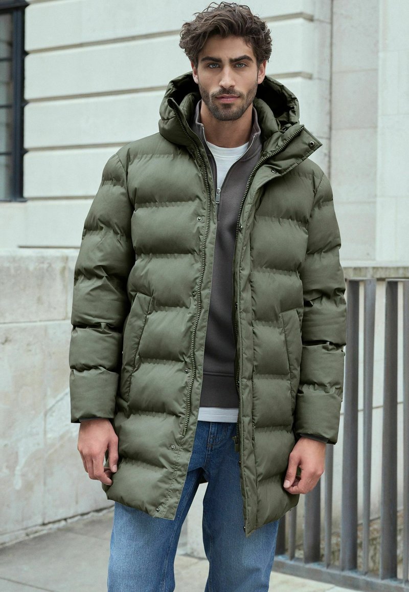 Polstret olivengrøn parka jakke med hætte, lynlås lukning, sidelommer og en glat tekstur, båret over en grå sweater og blå jeans.