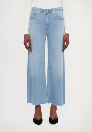 Lichtblauwe, wijduitlopende cropped spijkerbroeken met rafelige zoom, gedragen met zwarte pumps met spitse neus en een wit topje met lange mouwen.