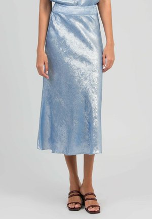 Femme portant une jupe midi bleu clair brillante avec une taille ajustée et des sandales à talons ouvertes marron, debout devant un fond uni.